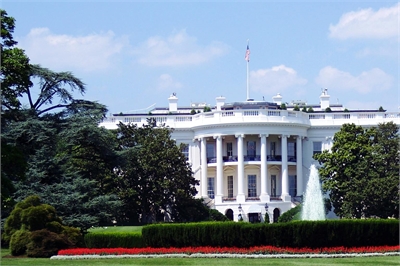 [ai] The White House seen from the front yard, featuring the iconic columns and a flag atop the roof, surrounded by green trees and well-maintained flower beds.